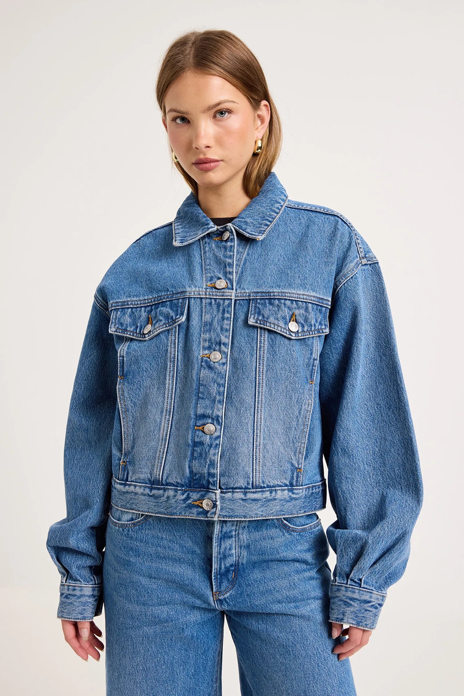 A woman modeling a cropped blue denim jacket with front flap pockets and button closure, paired with matching blue jeans, against a plain light gray studio background.