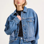 A woman modeling a cropped blue denim jacket over a black top and blue jeans, photographed against a plain light background.