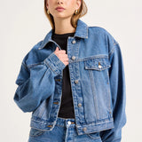A woman modeling a cropped blue denim jacket over a black top and blue jeans, photographed against a plain light background.