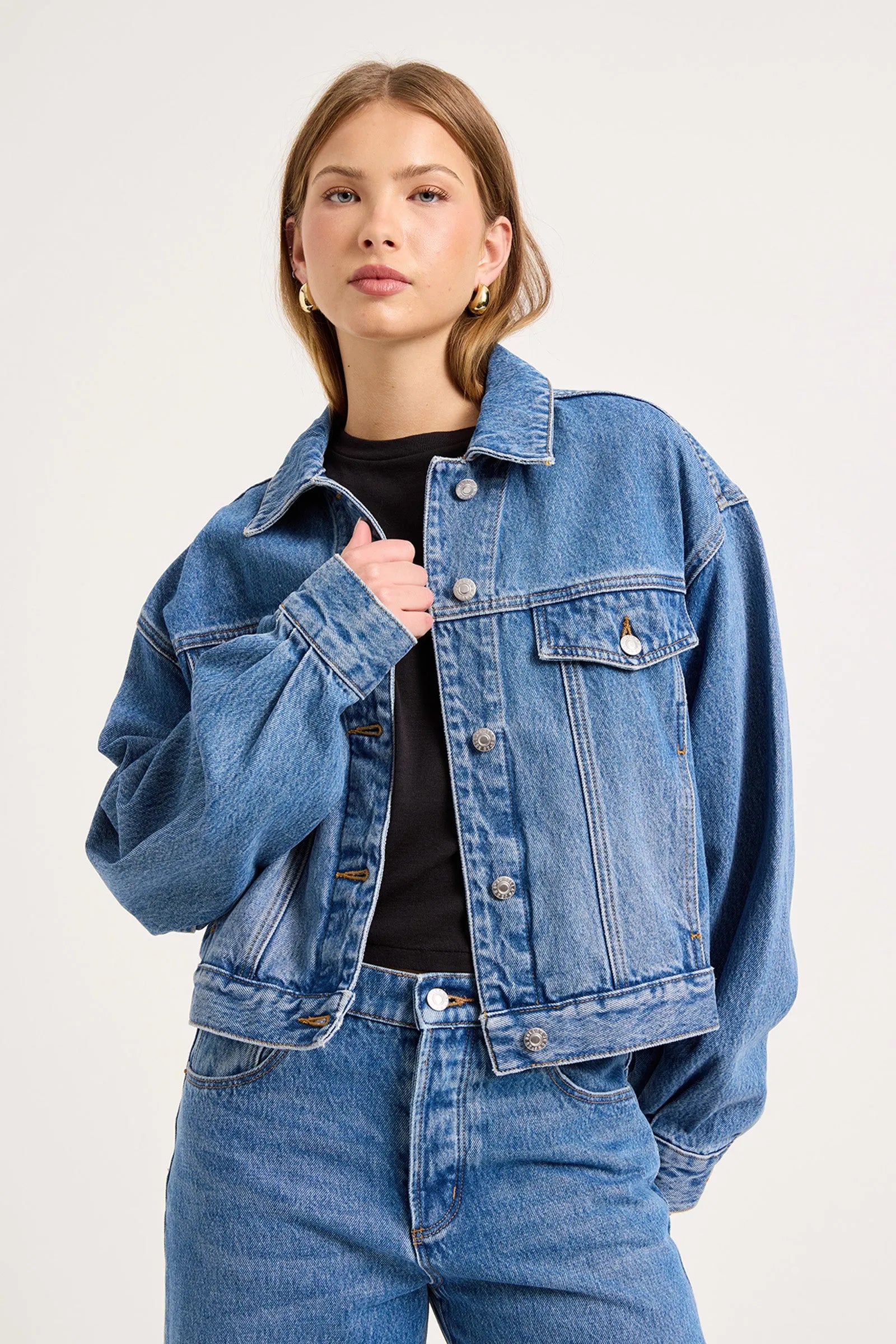 A woman modeling a cropped blue denim jacket over a black top and blue jeans, photographed against a plain light background.