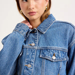 A close-up of a model wearing a classic blue denim jacket with silver buttons and a pointed collar, set against a plain light gray studio background.