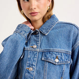 A close-up of a model wearing a classic blue denim jacket with silver buttons and a pointed collar, set against a plain light gray studio background.