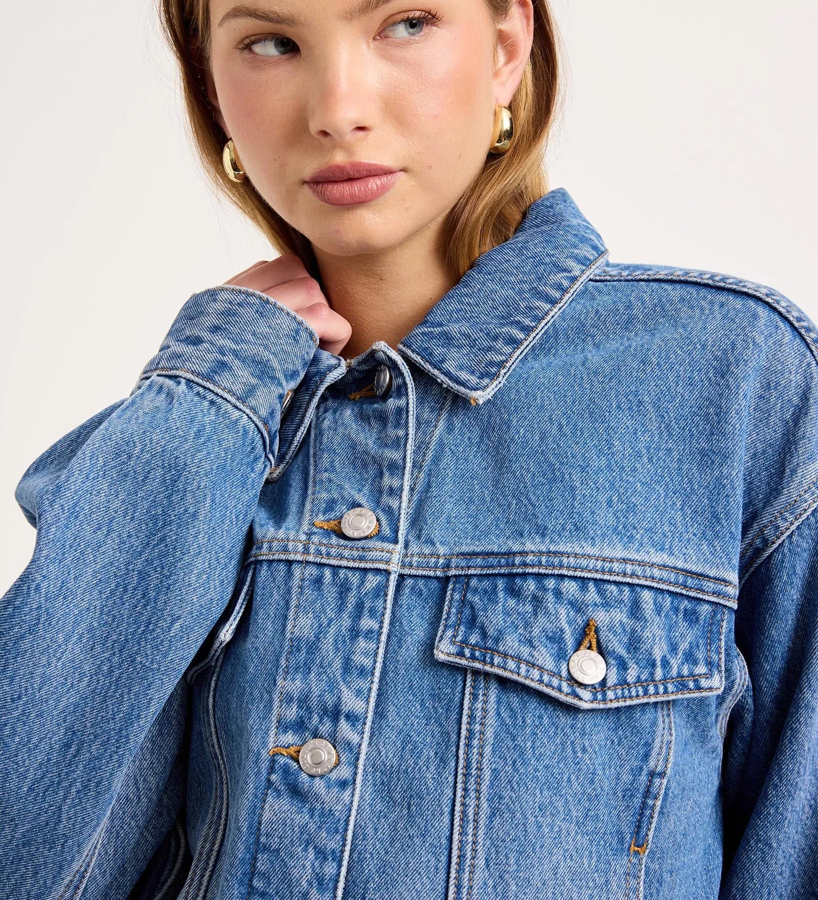 A close-up of a model wearing a classic blue denim jacket with silver buttons and a pointed collar, set against a plain light gray studio background.