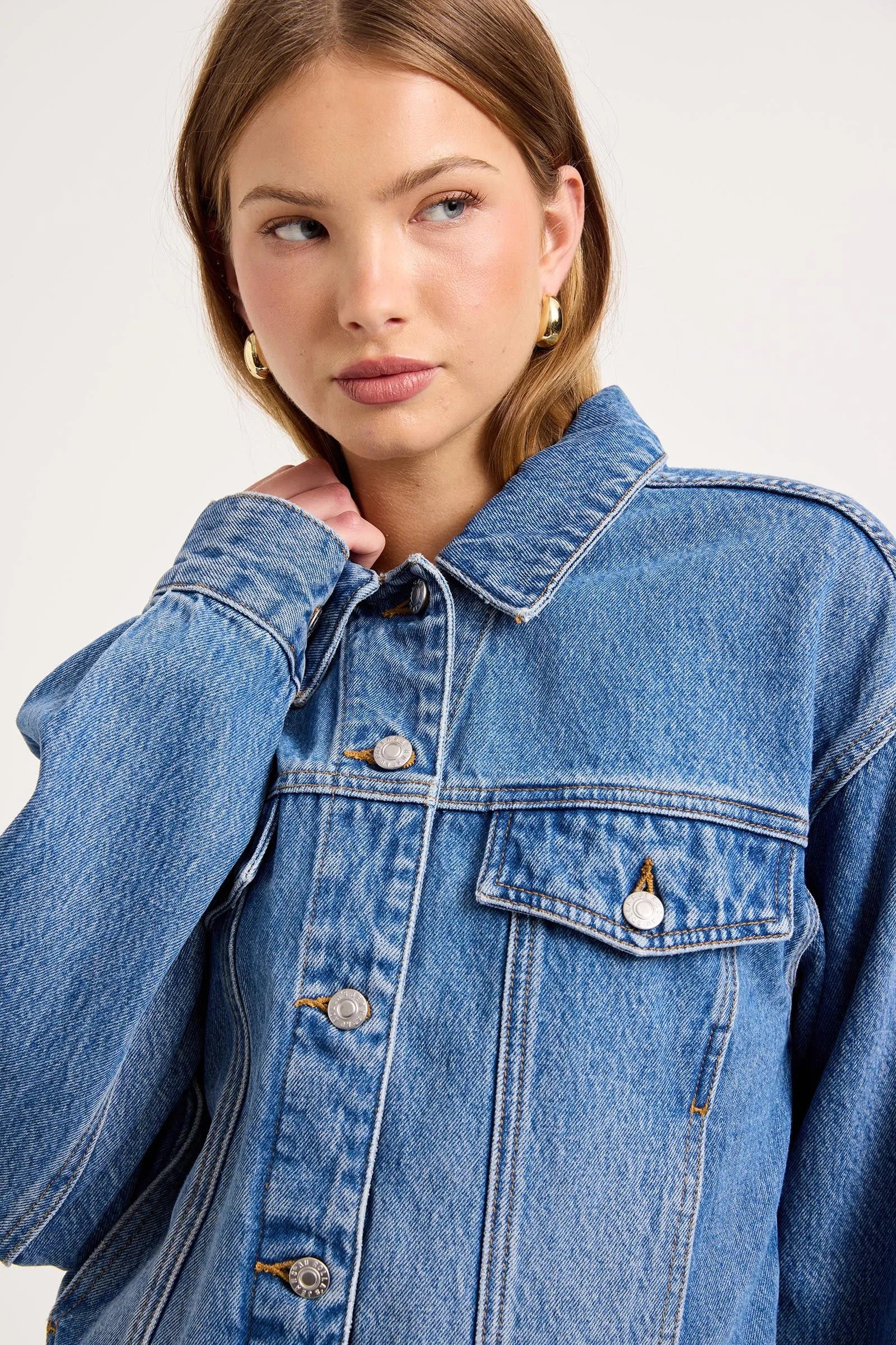 A close-up of a model wearing a classic blue denim jacket with silver buttons and a pointed collar, set against a plain light gray studio background.