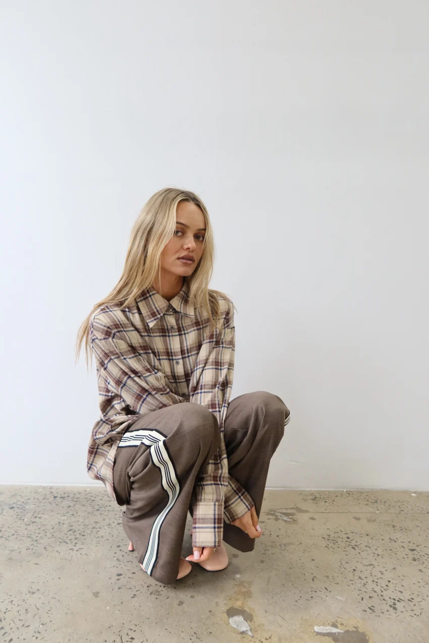 A woman crouching on a concrete floor against a plain white wall, wearing an oversized brown and beige plaid button-up shirt and matching brown sweatpants with white side stripes.