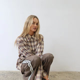 A woman crouching on a concrete floor against a plain white wall, wearing an oversized brown and beige plaid button-up shirt and matching brown sweatpants with white side stripes.