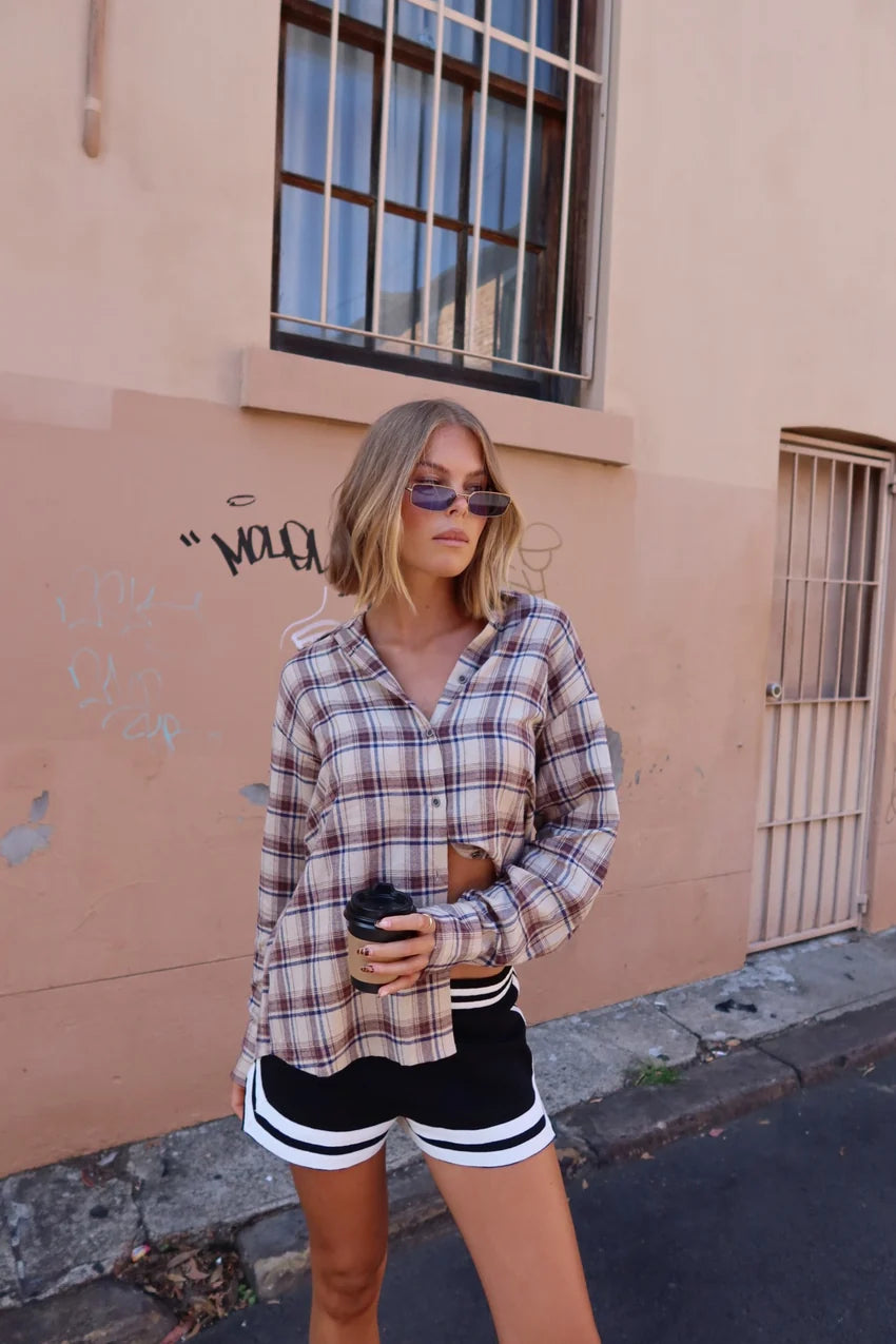 Woman in plaid shirt and shorts standing against a beige wall.