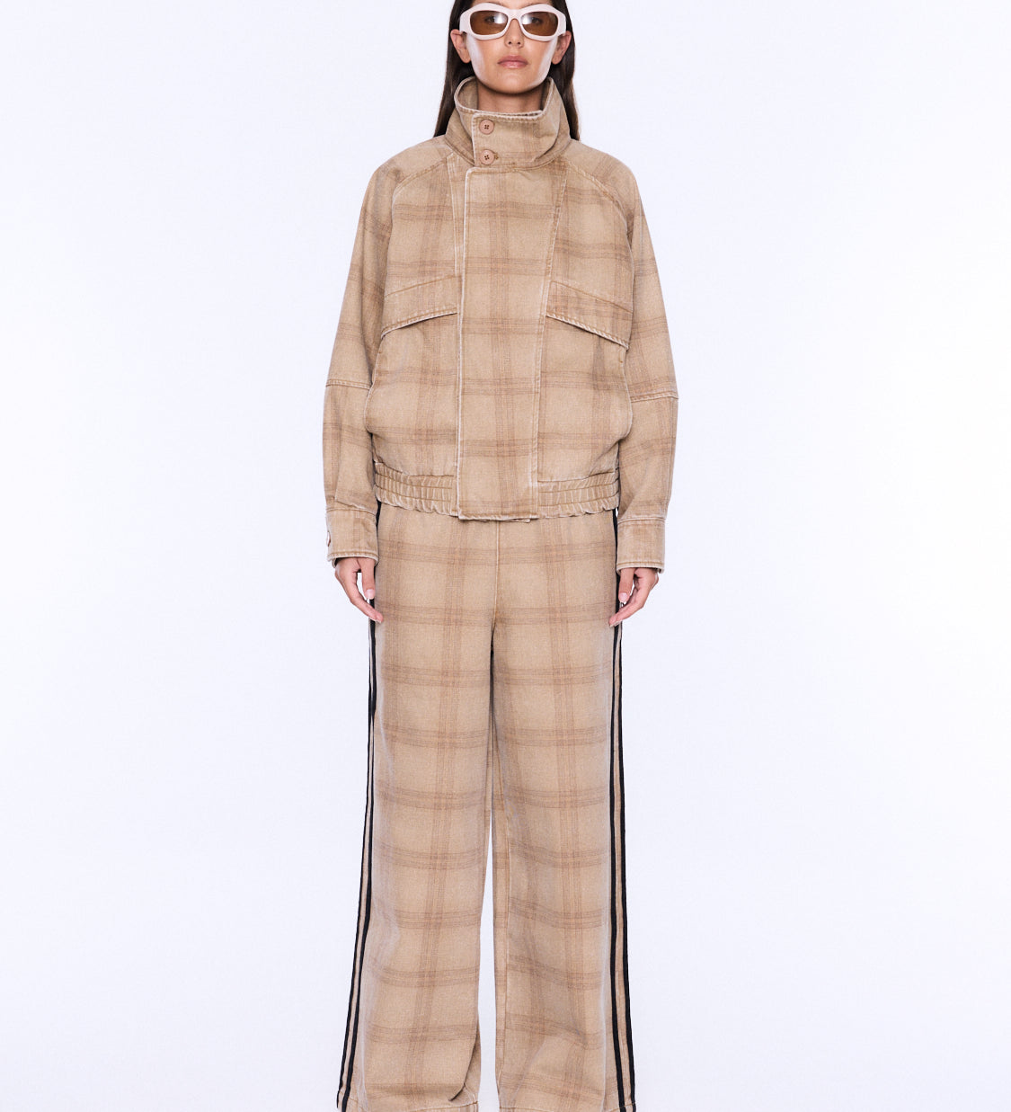 A model stands against a plain white studio background wearing a beige and tan plaid matching set consisting of a high-neck jacket and wide-leg trousers with black side stripes.