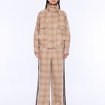 A model stands against a plain white studio background wearing a beige and tan plaid matching set consisting of a high-neck jacket and wide-leg trousers with black side stripes.