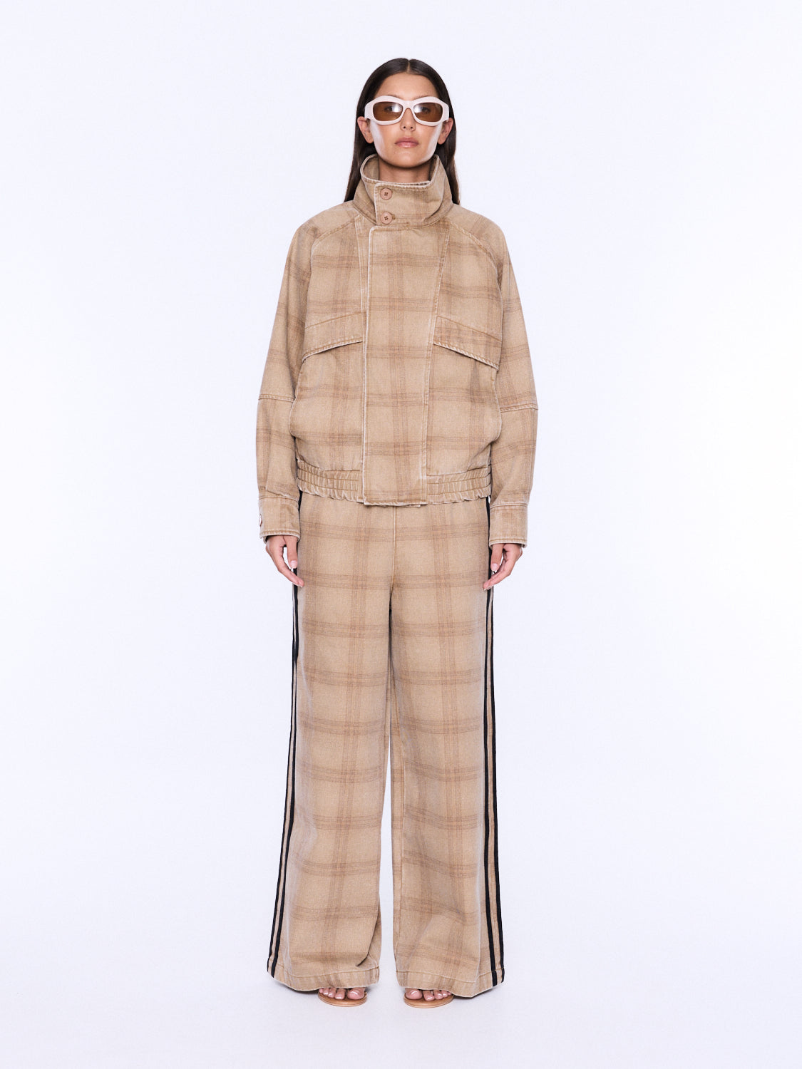 A model stands against a plain white studio background wearing a beige and tan plaid matching set consisting of a high-neck jacket and wide-leg trousers with black side stripes.