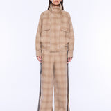 A model stands against a plain white studio background wearing a beige and tan plaid matching set consisting of a high-neck jacket and wide-leg trousers with black side stripes.