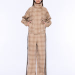 A model wearing a beige and brown plaid matching jacket and wide-leg pants set with black side stripes, styled with white-framed sunglasses against a plain white background.