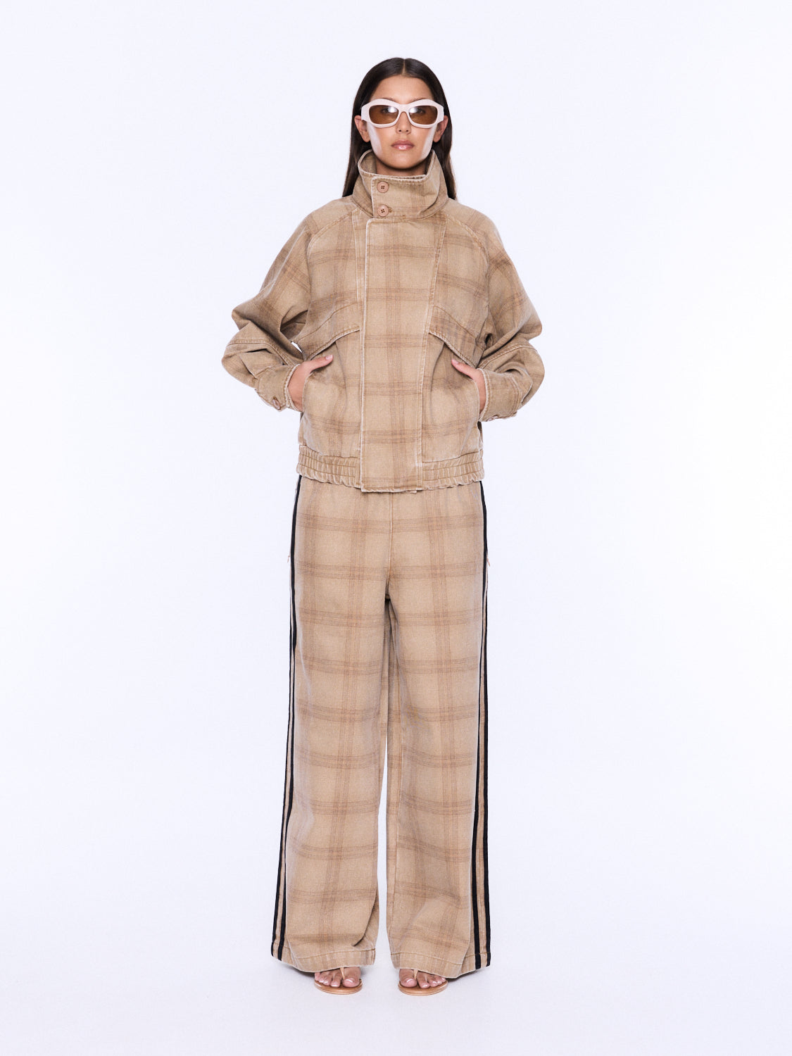 A model wearing a beige and brown plaid matching jacket and wide-leg pants set with black side stripes, styled with white-framed sunglasses against a plain white background.