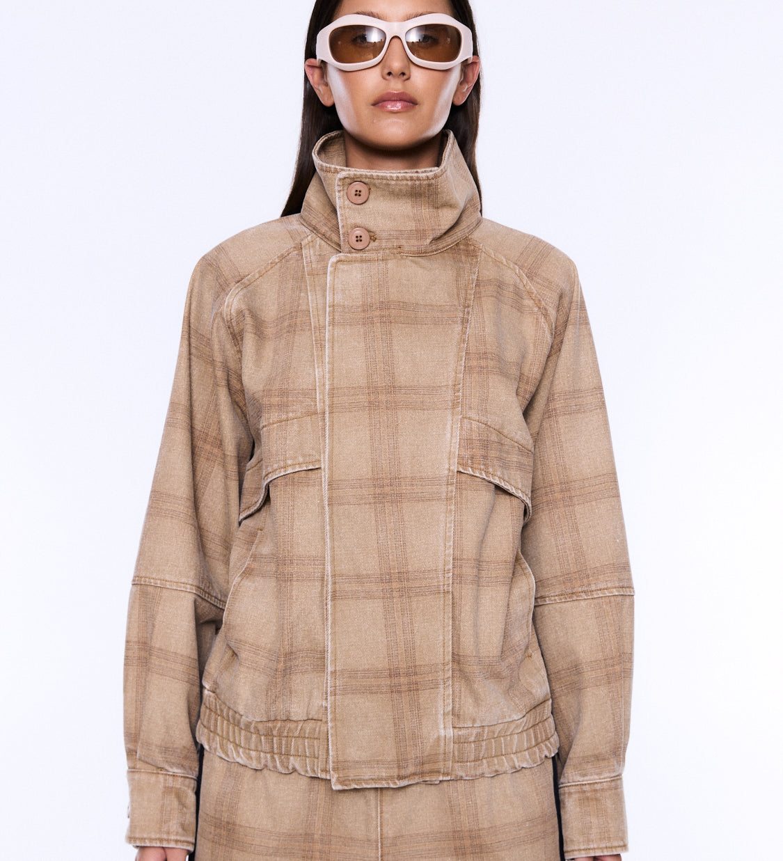 A model wearing a beige and brown plaid jacket with a high mock neck and button closure, paired with matching plaid pants and white-framed sunglasses, against a plain white background.
