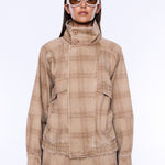 A model wearing a beige and brown plaid jacket with a high mock neck and button closure, paired with matching plaid pants and white-framed sunglasses, against a plain white background.