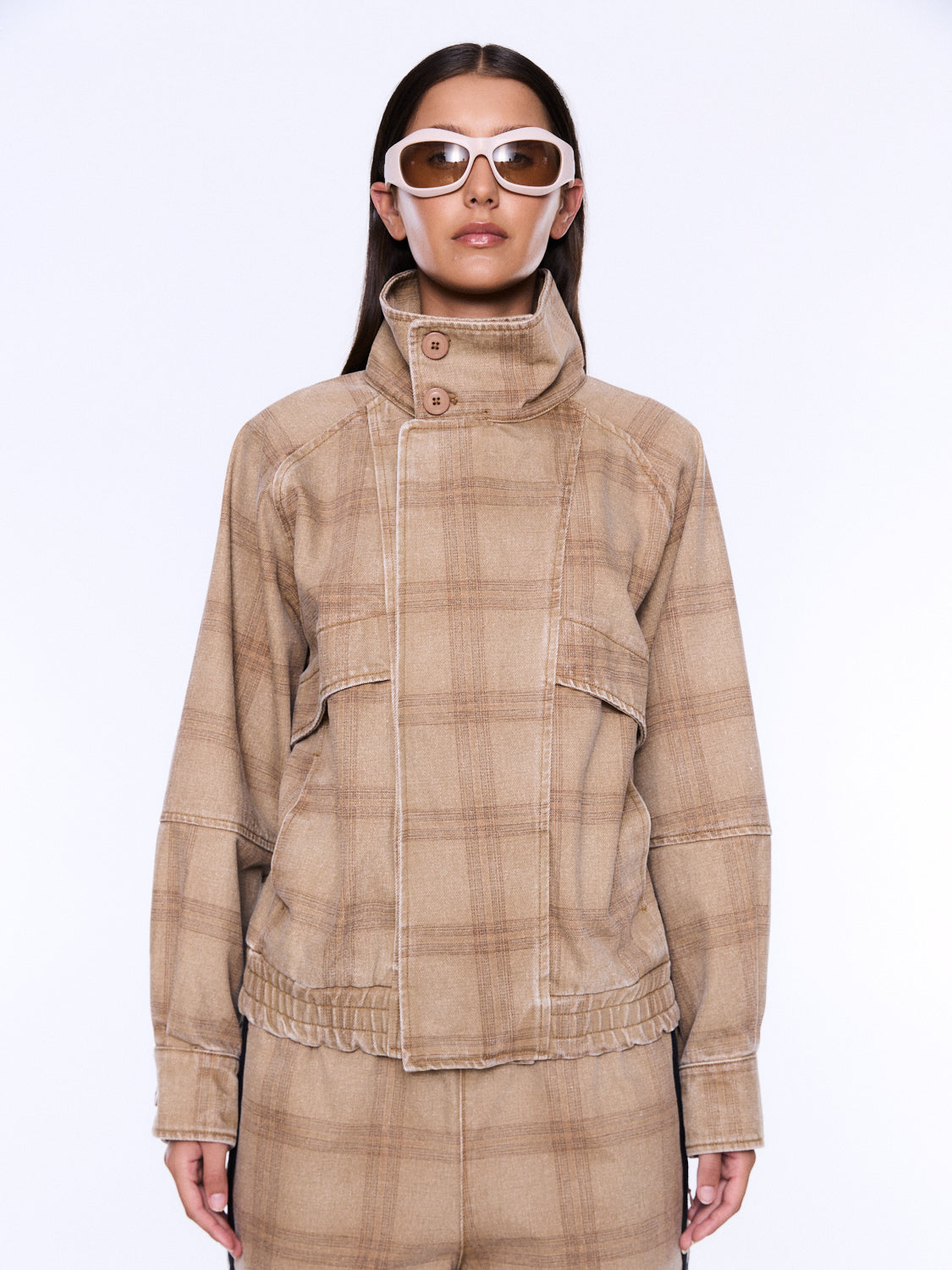 A model wearing a beige and brown plaid jacket with a high mock neck and button closure, paired with matching plaid pants and white-framed sunglasses, against a plain white background.