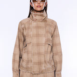 A model wearing a beige and brown plaid jacket with a high mock neck and button closure, paired with matching plaid pants and white-framed sunglasses, against a plain white background.