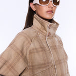 A close-up of a model wearing a beige and brown plaid jacket with a high collar and white-framed sunglasses against a plain white background.