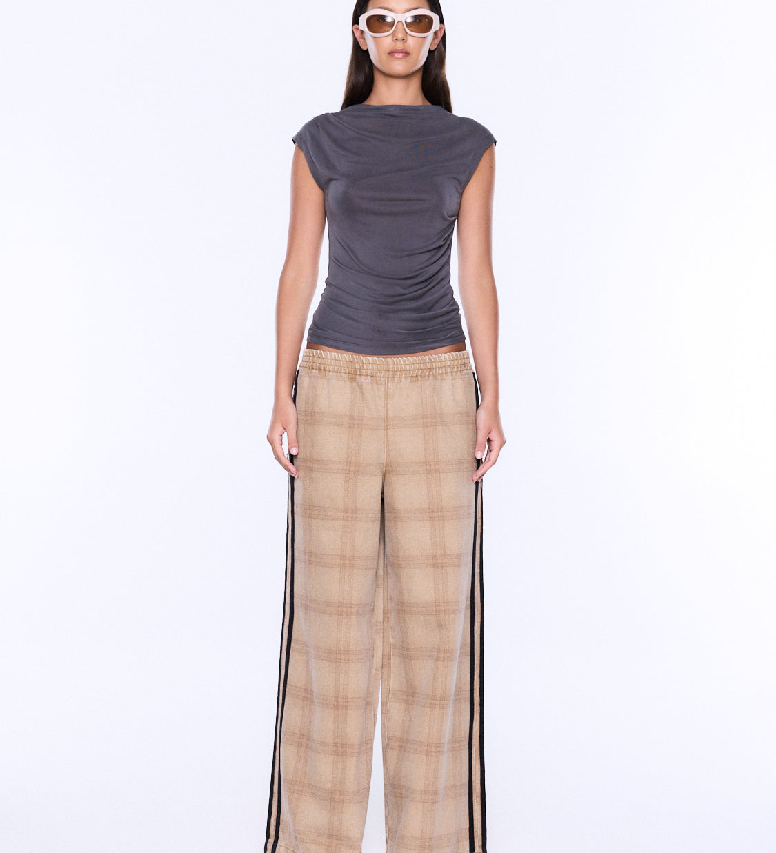 A model stands against a plain white studio background wearing a dark gray sleeveless top and beige plaid wide-leg pants with black side stripes.