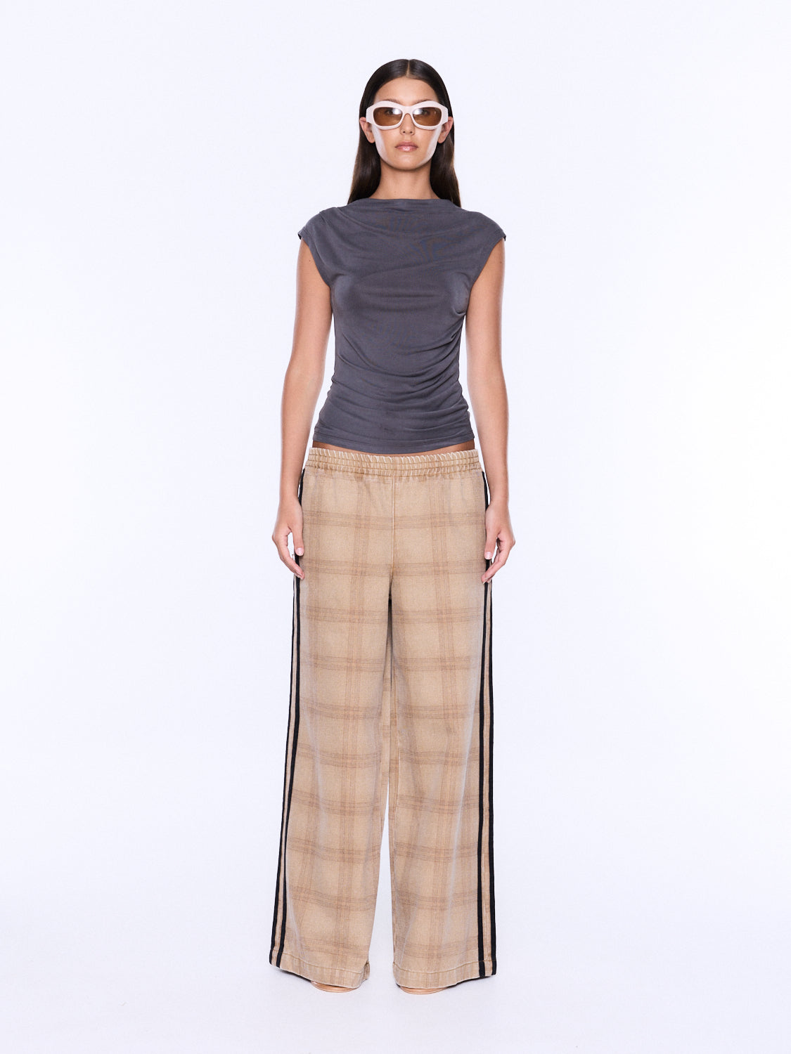 A model stands against a plain white studio background wearing a dark gray sleeveless top and beige plaid wide-leg pants with black side stripes.