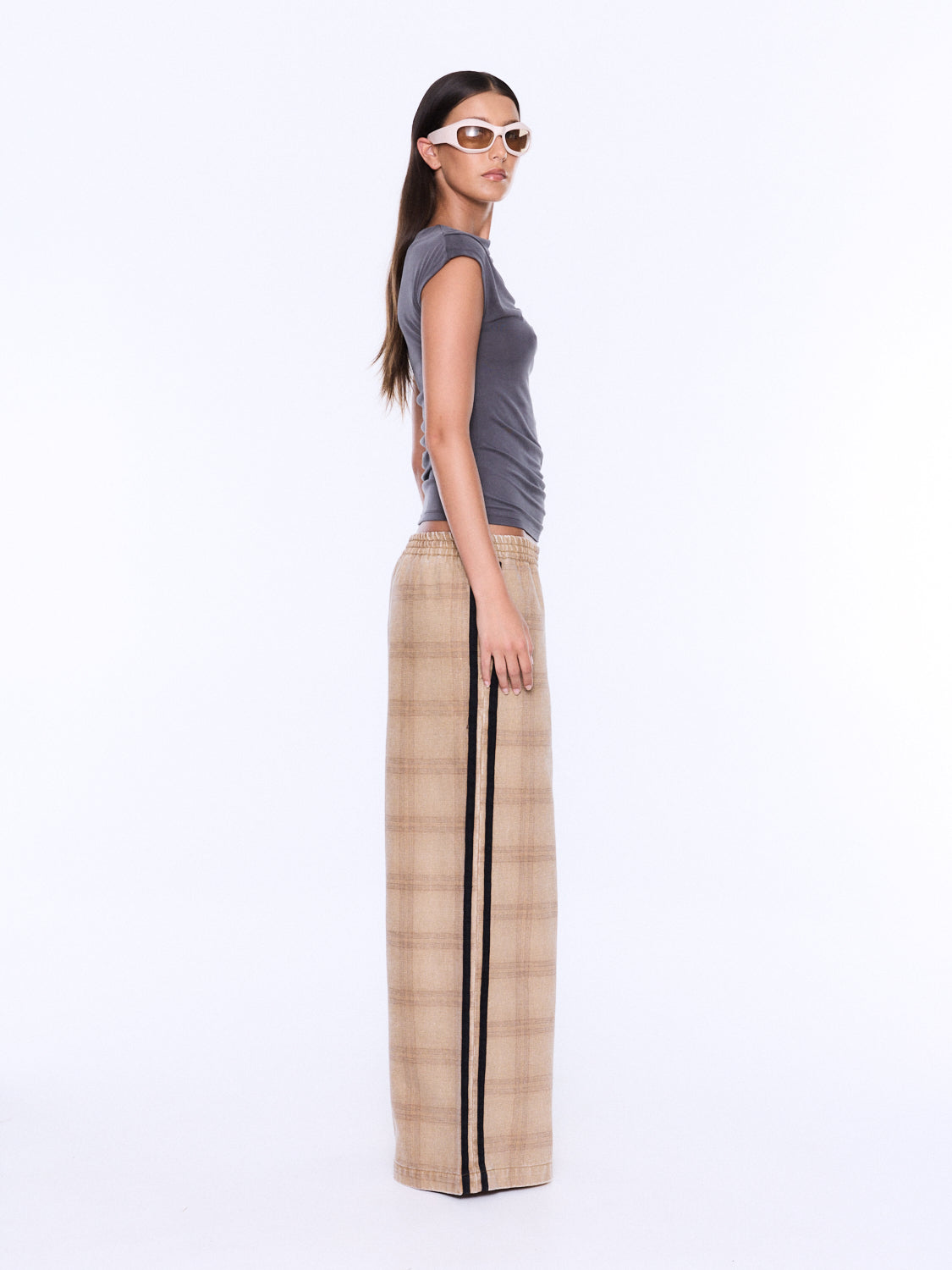 A woman stands in profile wearing a gray sleeveless top and beige plaid wide-leg pants with a black side stripe, photographed against a plain white studio background.