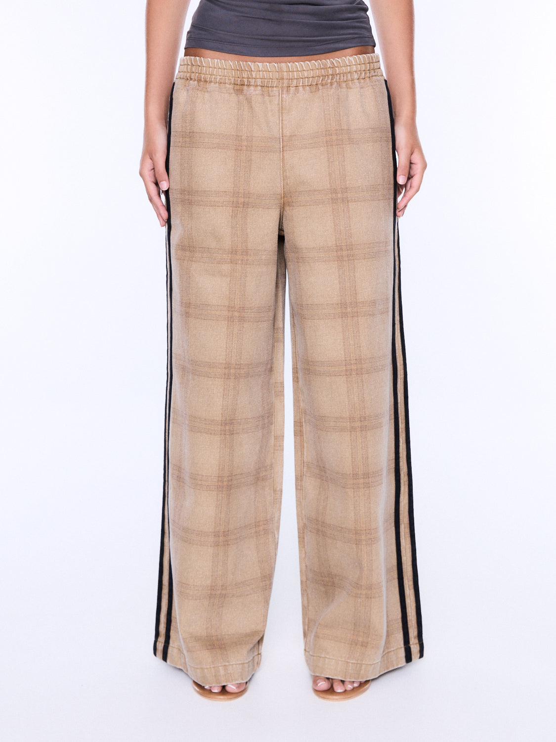 A person wearing wide-leg plaid pants in beige and brown with black side stripes, paired with a dark gray top and tan sandals, shown against a plain white studio background.