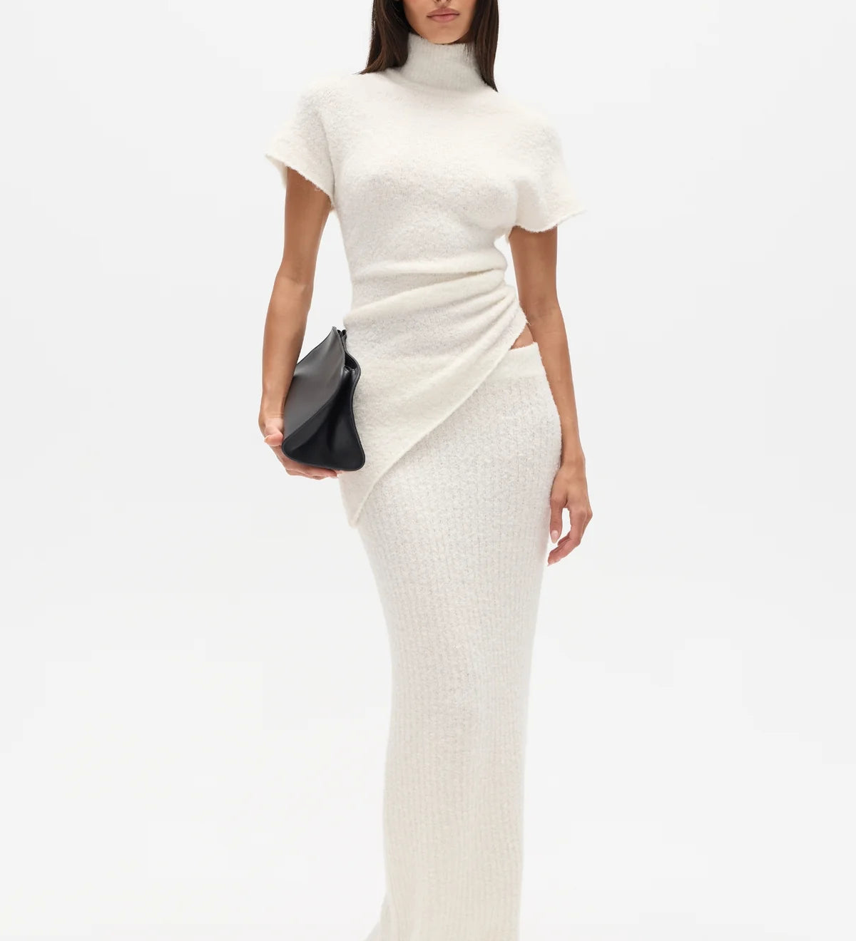 Woman wearing a white high neck top and long white skirt with a black clutch on a white background