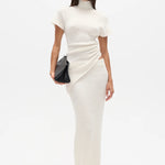 Woman wearing a white high neck top and long white skirt with a black clutch on a white background