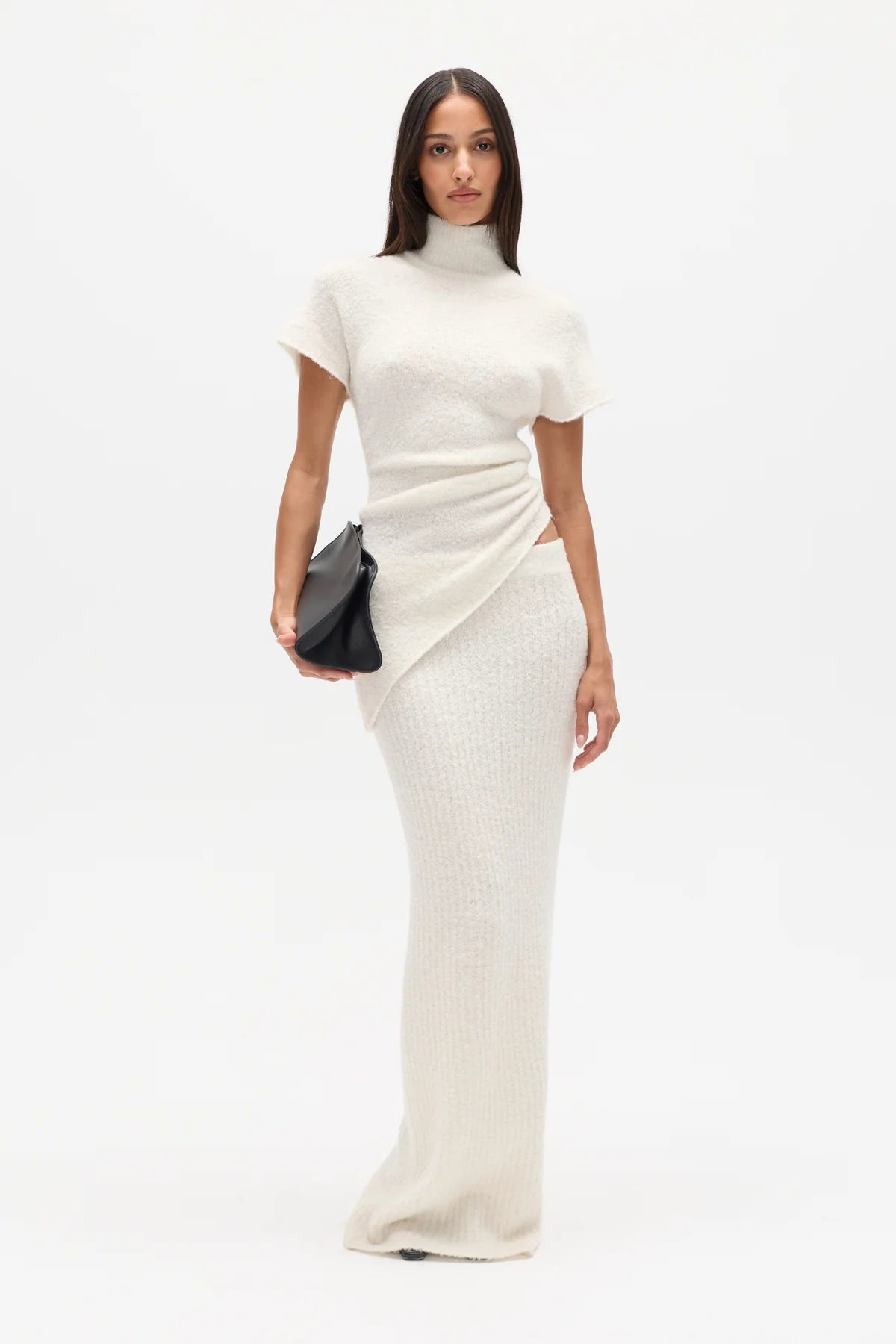 Woman wearing a white high neck top and long white skirt with a black clutch on a white background