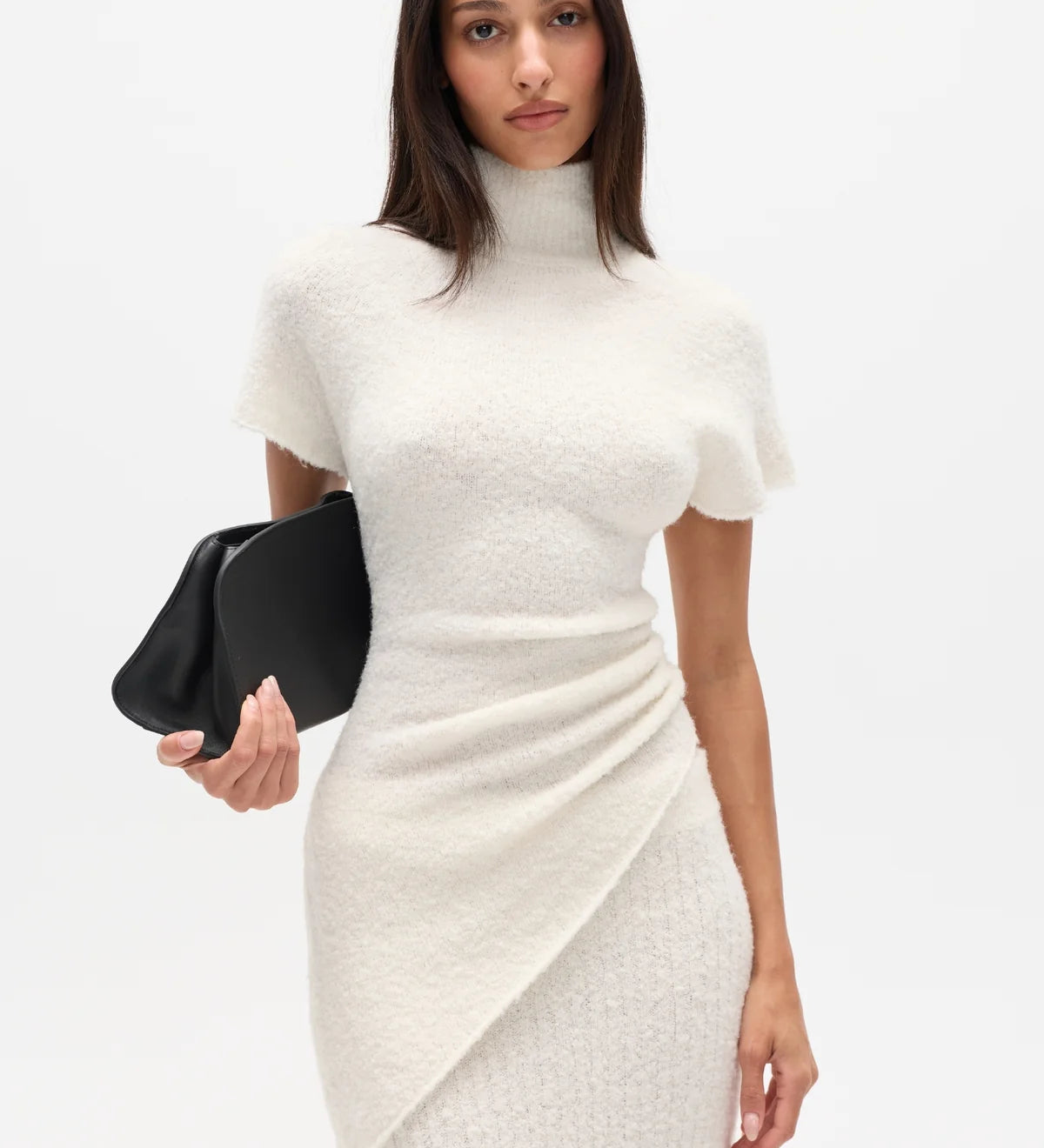 Woman wearing a white high neck top and long white skirt with a black clutch on a white background