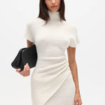 Woman wearing a white high neck top and long white skirt with a black clutch on a white background