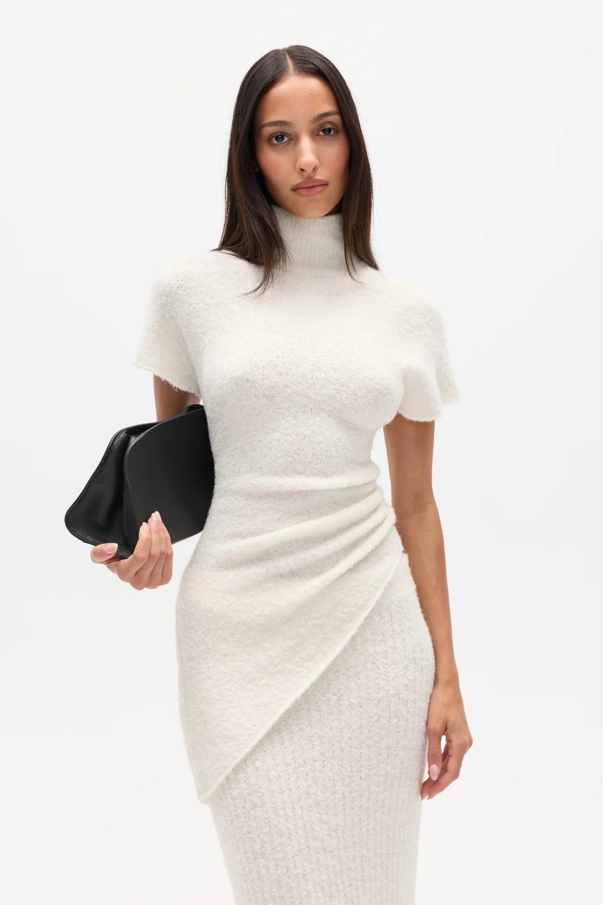 Woman wearing a white high neck top and long white skirt with a black clutch on a white background