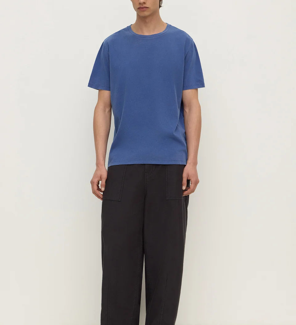 A man wearing a blue short-sleeve crewneck t-shirt and loose black trousers, standing against a plain off-white studio background.