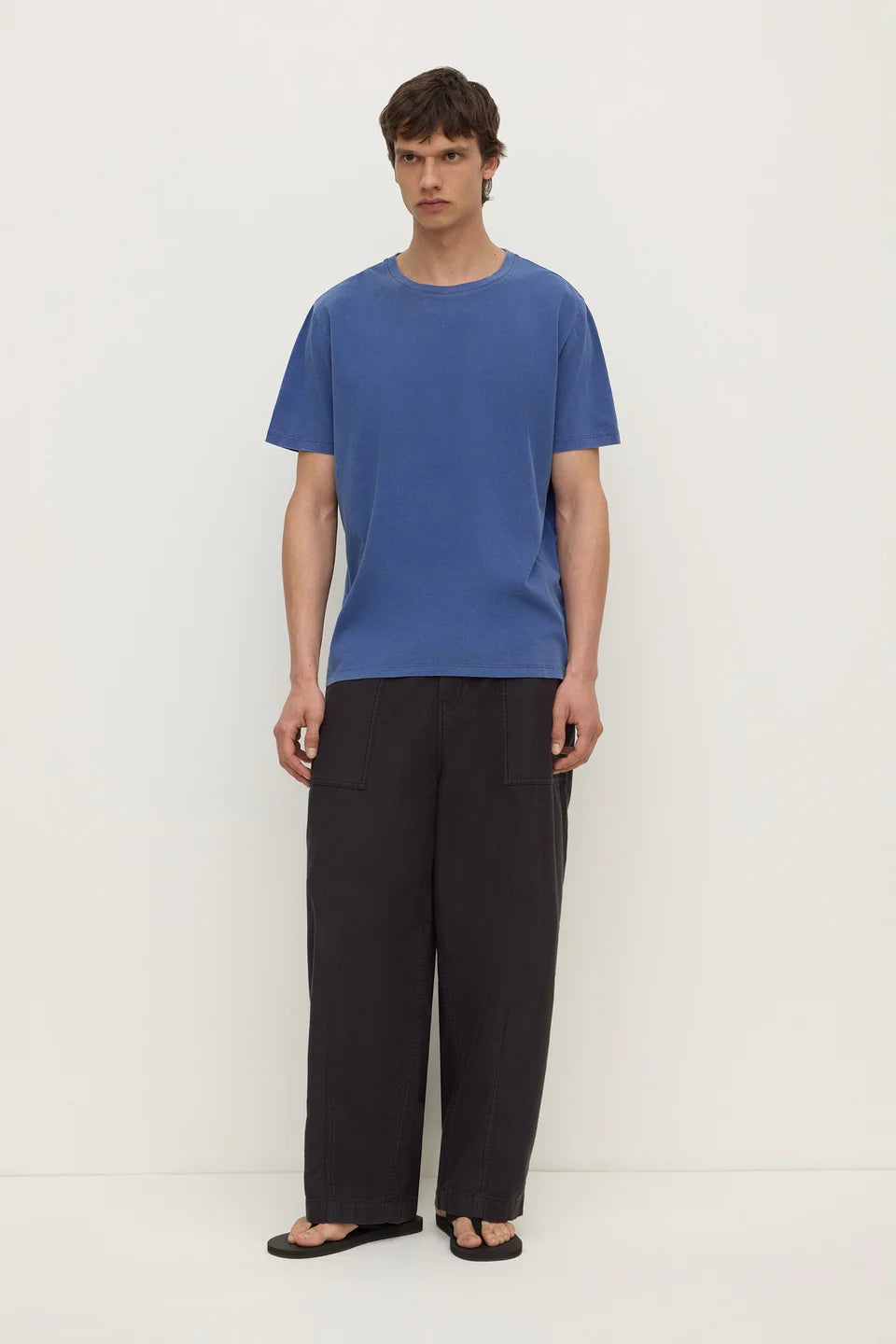 A man wearing a blue short-sleeve crewneck t-shirt and loose black trousers, standing against a plain off-white studio background.