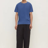 A man wearing a blue short-sleeve crewneck t-shirt and loose black trousers, standing against a plain off-white studio background.
