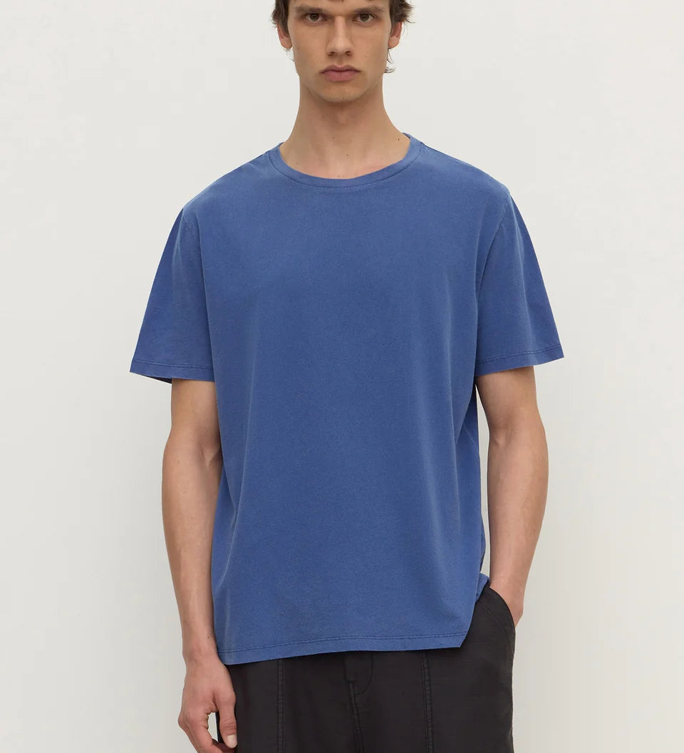 A man wearing a blue short-sleeve crewneck t-shirt and dark pants, standing against a plain off-white background.