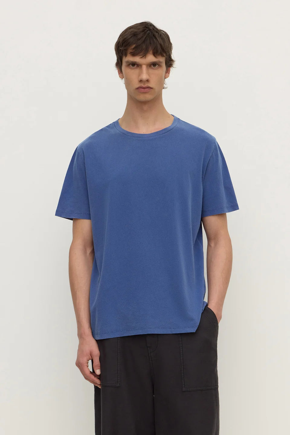 A man wearing a blue short-sleeve crewneck t-shirt and dark pants, standing against a plain off-white background.