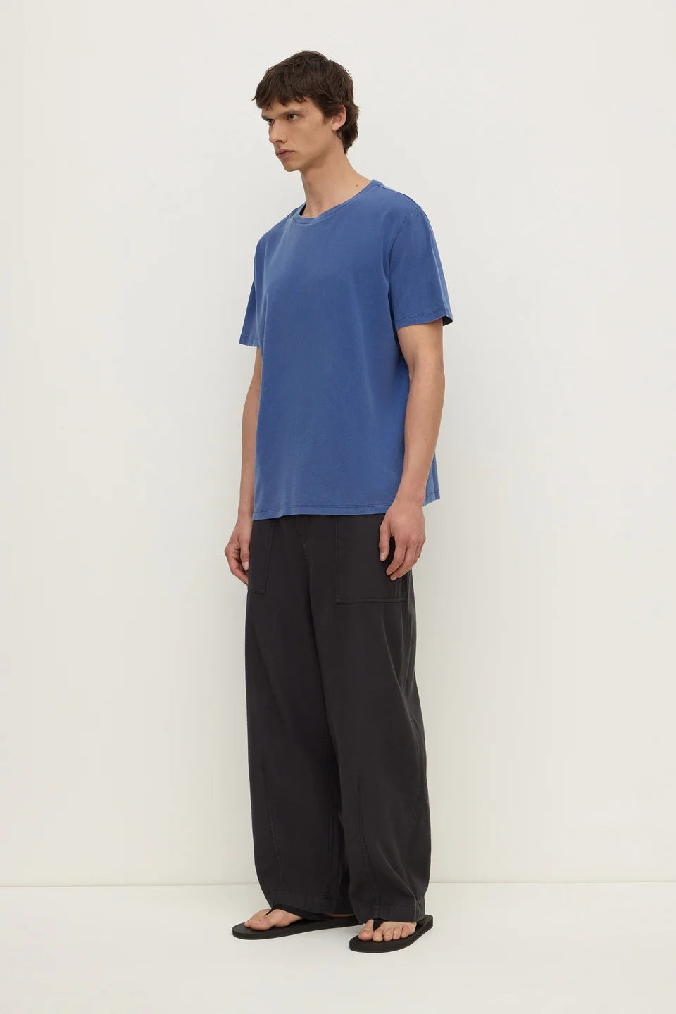 A man wearing a blue short-sleeve crewneck t-shirt, loose black wide-leg pants, and black sandals, standing against a plain off-white studio background.