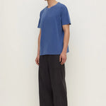 A man wearing a blue short-sleeve crewneck t-shirt, loose black wide-leg pants, and black sandals, standing against a plain off-white studio background.