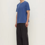A man wearing a blue short-sleeve crewneck t-shirt, loose black wide-leg pants, and black sandals, standing against a plain off-white studio background.