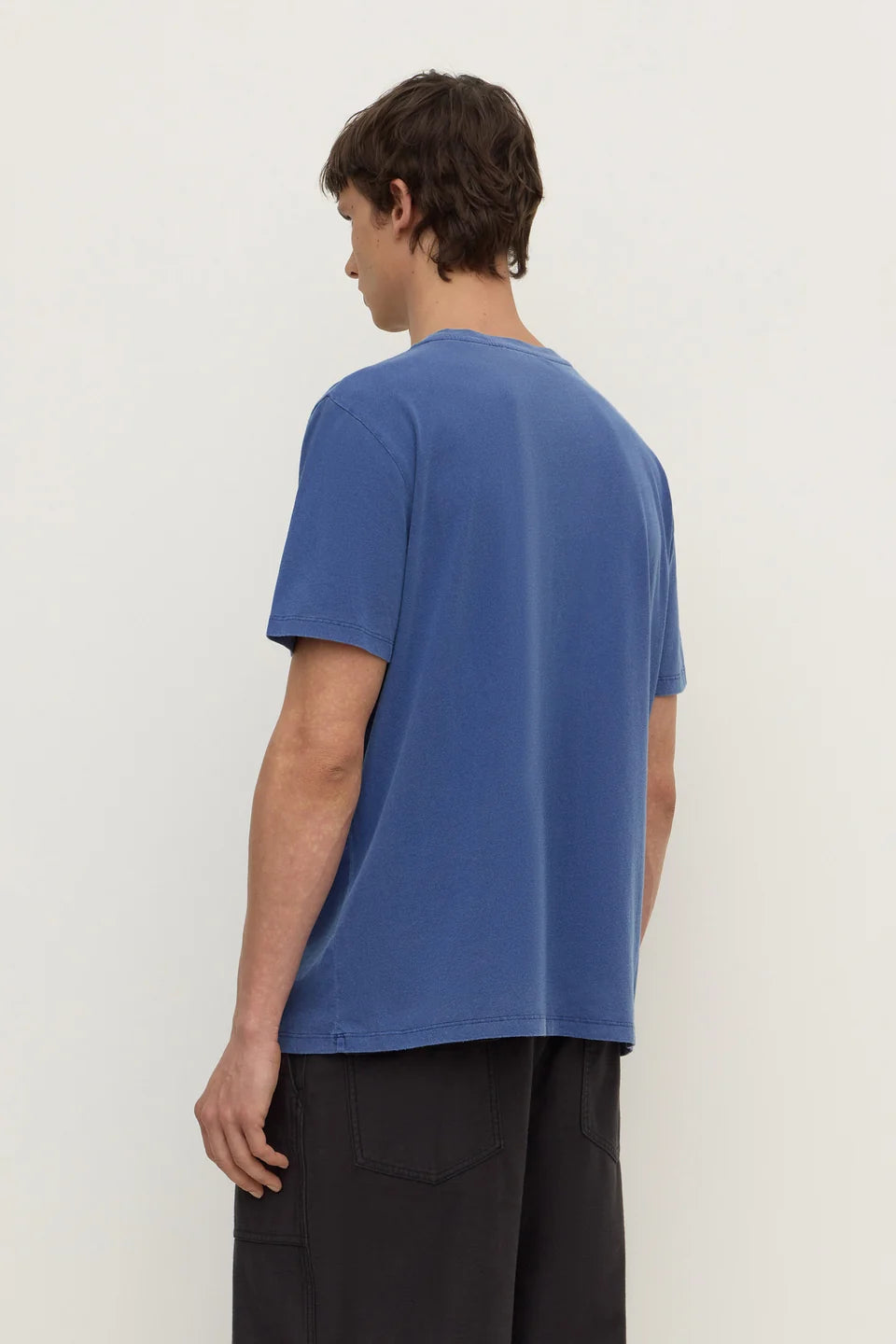 A man wearing a blue short-sleeve t-shirt and dark pants, shown from the back against a plain off-white studio background.