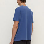 A man wearing a blue short-sleeve t-shirt and dark pants, shown from the back against a plain off-white studio background.