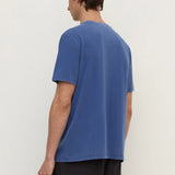 A man wearing a blue short-sleeve t-shirt and dark pants, shown from the back against a plain off-white studio background.
