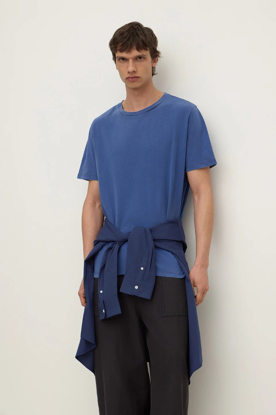 A male model wearing a blue short-sleeve crewneck t-shirt with a matching blue shirt tied around the waist as a belt, paired with dark gray trousers, against a plain off-white background.