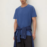 A male model wearing a blue short-sleeve crewneck t-shirt with a matching blue shirt tied around the waist as a belt, paired with dark gray trousers, against a plain off-white background.