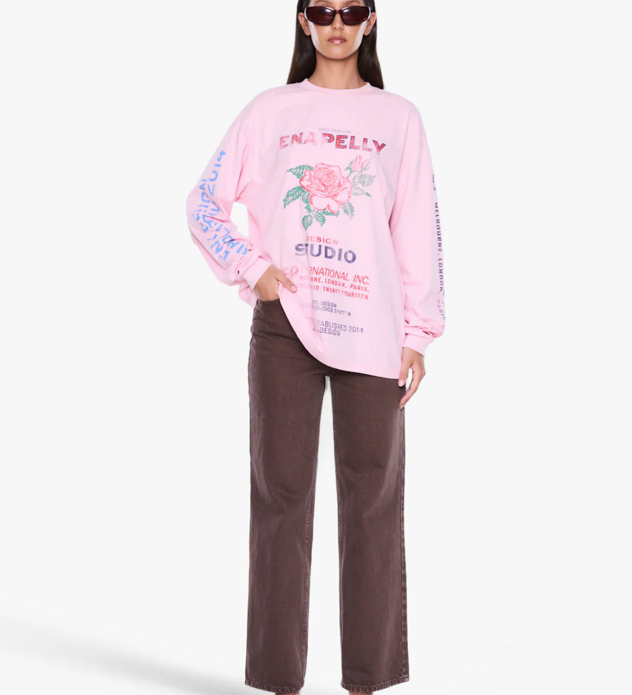 A woman modeling a light pink long-sleeve graphic sweatshirt with a floral print and text, paired with dark brown wide-leg pants, against a plain light gray studio background.