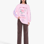 A woman modeling a light pink long-sleeve graphic sweatshirt with a floral print and text, paired with dark brown wide-leg pants, against a plain light gray studio background.