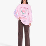 A woman modeling a light pink long-sleeve graphic sweatshirt with a floral print and text, paired with dark brown wide-leg pants, against a plain light gray studio background.