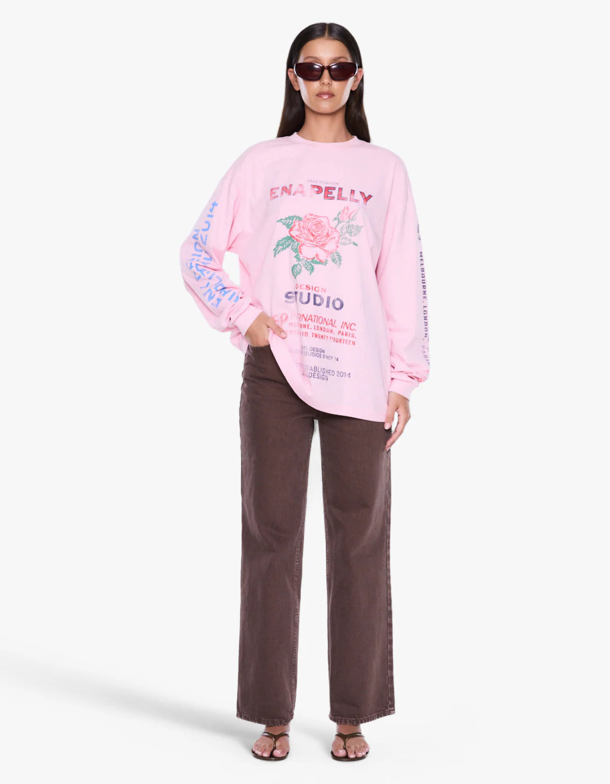 A woman modeling a light pink long-sleeve graphic sweatshirt with a floral print and text, paired with dark brown wide-leg pants, against a plain light gray studio background.