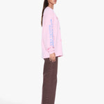 A woman modeling a light pink long-sleeve t-shirt with vertical blue text on the sleeve, paired with brown wide-leg pants and sunglasses, shown from a side profile against a light gray background.
